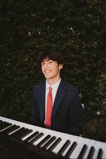 Jake Weiss smiling while sitting at a piano with a beautiful dark green bush behind him.
